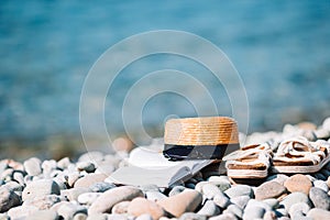 Beach hat on opened book with sunscreen and shoes on pebble beach