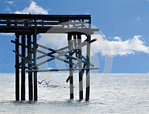 The beach fishing pier has a flock of pelicans fly by the end of the wood structure