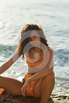 Beach Girl Model in Orange Bikini on the Rocky Deserted Seashore During Summer Beach Holidays