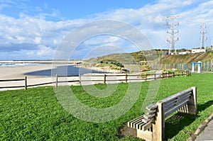 Beach with grass, wooden bench and fence. Industrial port and blue river, cloudy sky. Sabon, Galicia, Spain.