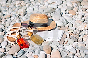 Beach hat on opened book with sunscreen and shoes on pebble beach