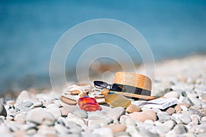 Beach hat on opened book with sunscreen and shoes on pebble beach