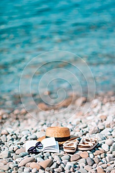 Beach hat on opened book with sunscreen and shoes on pebble beach