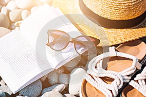 Beach hat on opened book with sunscreen and shoes on pebble beach