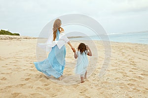 Beach. Kid And Woman In Maxi Dress Walking Along Ocean Coast. View From Back Of Young Mother With Daughter At Resort.