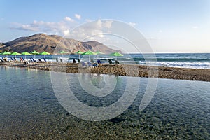 Beach lounge chairs under umbrellas on sandy peninsula between sea and river near Georgioupoli, Crete