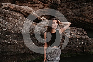 A beach photo of a beautiful sensual young woman with long dark hair in a dress posing on the beach by large boulders.