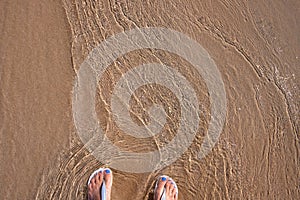 Beach sand and woman feet in sea wave top view. Relaxing photo of sandy beach and clean seawater. Girl feet in sandals