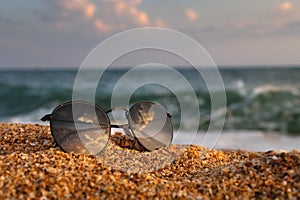 On the beach, sunglasses lie on the sand in the background a wave rolls from the sea. Close-up of leisure accessory, no
