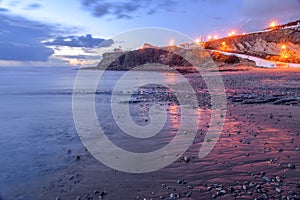 Beach surrounded by the sea and cliffs covered in lamps under a cloudy sky in the evening