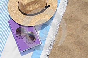 Beach towel with book, sunglasses and straw hat on sand, flat lay. Space for text