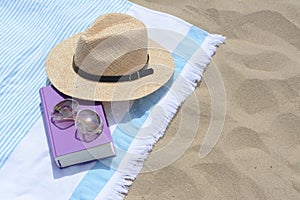 Beach towel with book, sunglasses and straw hat on sand, space for text