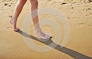 Beach travel - young girl walking on sand beach leaving footprints in the sand. Closeup detail of female feet and golden sand