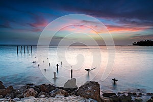 Beach at twilight. Exposed pier pylons extending into the ocean. Silky water. Pink clouds in sky above.