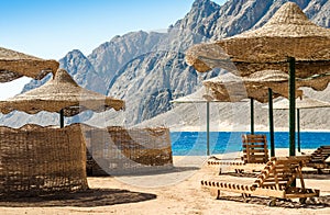 Beach umbrellas and wooden lounge chairs on the sand of the beach against the backdrop of the sea and high rocky mountains in