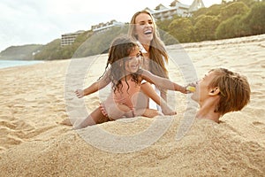 Beach Vacation. Family Enjoying Summertime At Tropical Resort. Little Sister Feeds Her Buried In Sand Brother Fruit.