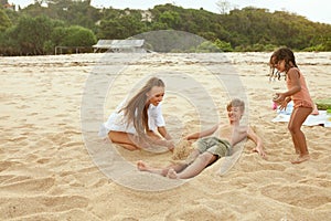 Beach Vacation. Family Enjoying Summertime At Tropical Resort. Mother And Little Girl Burying Boy In Sand.