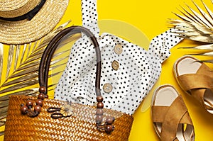 Beach wicker straw or rattan women`s eco bag, white dress straw hat sandals golden tropical leaf on yellow background Flat lay to