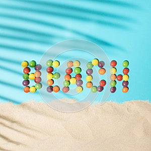Beach word made from colored balls on a blue background with palm leaf shadow and sand. Tropical summer flat lay beach concept