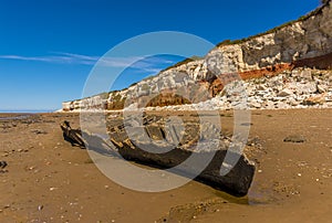 A beached carcass of a shipwreck on Old Hunstanton Beach beneath the white, red and orange stratified cliffs in Norfolk, UK