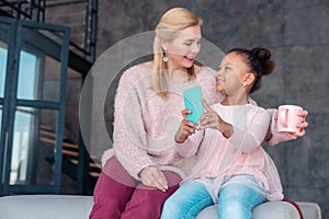 Beaming appealing girl holding blue smartphone sitting near mom