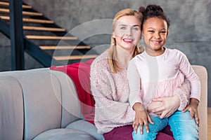 Beaming daughter sitting on knees of her lovely mother