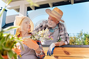 Beaming elderly lady holding white flowers looking at her handsome husband