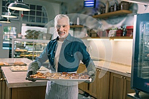 Beaming grey-haired baker holding whole tray with nice cinnamon buns