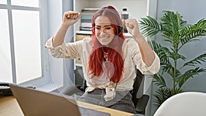 Beaming redhead business woman celebrating work victory indoors, young professional elatedly using laptop and headphones in office photo