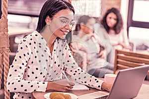 Beaming woman wearing spotted blouse looking at her laptop reading message