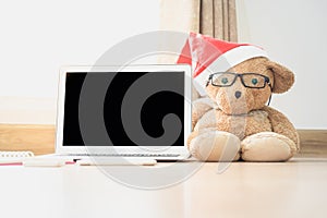 Bear doll wearing red hat sit beside laptop