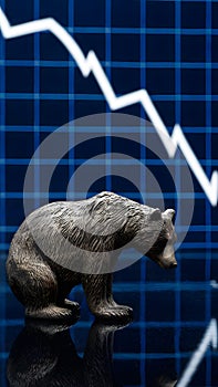 Bear figurine on reflective surface next to descending stock market graph, reflecting downturn sentiment