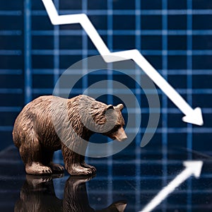 Bear figurine on reflective surface next to descending stock market graph, reflecting downturn sentiment
