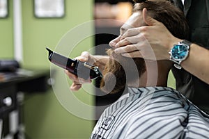 Beard grooming. Side view of young bearded man getting beard haircut by hairdresser while sitting in chair at barbershop