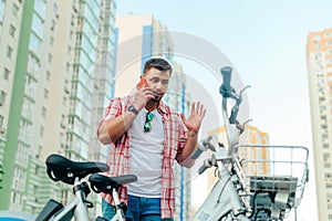 Bearded adult stands by a bicycle against the backdrop of buildings and the sky, talking on a smartphone with a serious face.