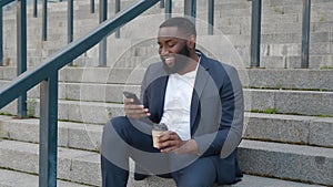 Bearded African American man browses the Internet on a modern smartphone and drinks coffee while sitting on the steps