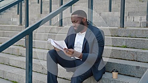 Bearded African American man looks through paper documents with business charts while sitting on the steps near the