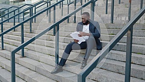 Bearded African American man looks through paper documents with business charts while sitting on the steps near the