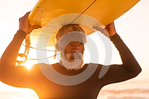 Bearded african american senior man carrying surfboard on head at beach against clear sky at sunset