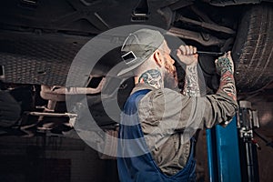 Bearded auto mechanic in a uniform repair the car`s suspension with a wrench while standing under lifting car in repair