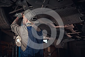 Bearded auto mechanic in a uniform repair the car`s suspension with a wrench while standing under lifting car in repair