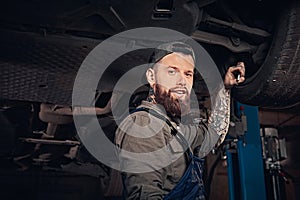 Bearded auto mechanic in a uniform repair the car`s suspension with a wrench while standing under lifting car in repair