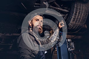 Bearded auto mechanic in a uniform repair the car`s suspension with a wrench while standing under lifting car in repair