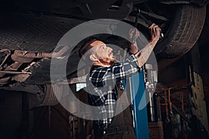 Bearded auto mechanic in a uniform repair the car`s suspension with a wrench while standing under lifting car in repair