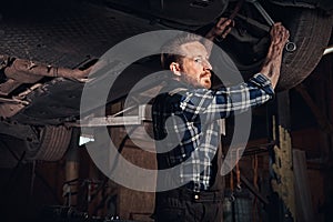 Bearded auto mechanic in a uniform repair the car`s suspension with a wrench while standing under lifting car in repair