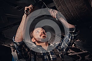 Bearded auto mechanic in a uniform repair the car`s suspension with a wrench while standing under lifting car in repair