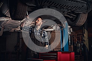 Bearded auto mechanic in a uniform repair the car`s suspension with a wrench while standing under lifting car in repair