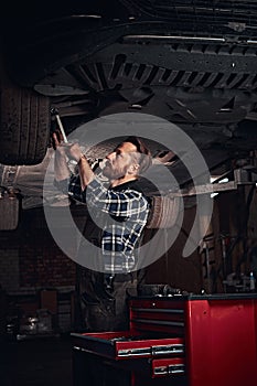 Bearded auto mechanic in a uniform repair the car`s suspension with a wrench while standing under lifting car in repair