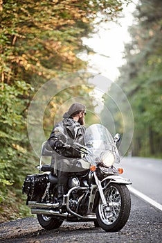 Bearded biker in black leather jacket on modern motorcycle on country roadside.