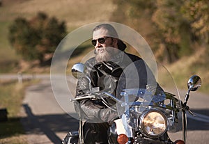 Bearded biker with long hair in black leather jacket and sunglasses sitting on modern motorcycle.